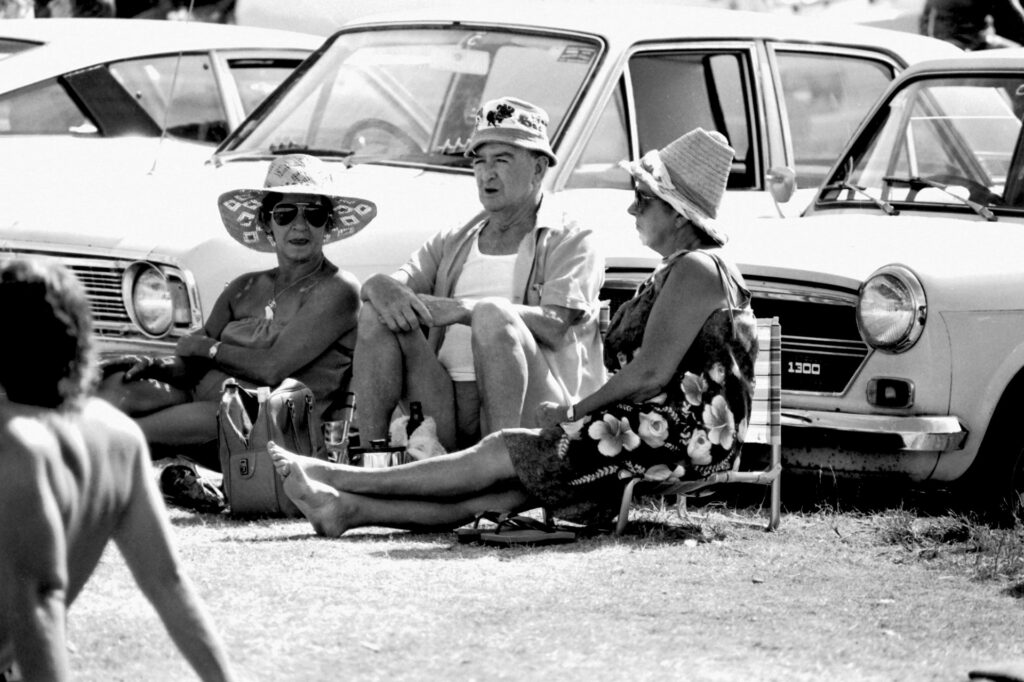 Personas sentadas en sillas plegables delante de coches aparcados. Blanco y negro.